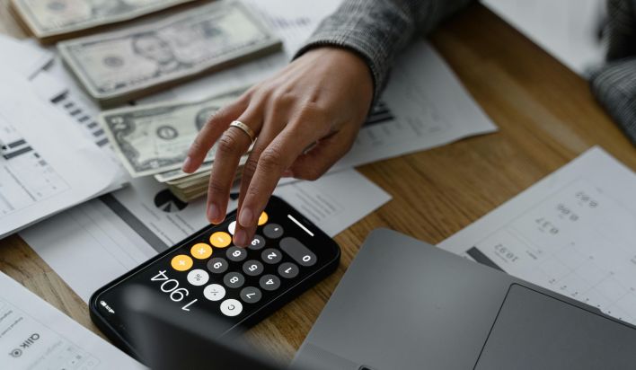 Hand of a Person Using a Calculator Near Cash Money on Wooden Table